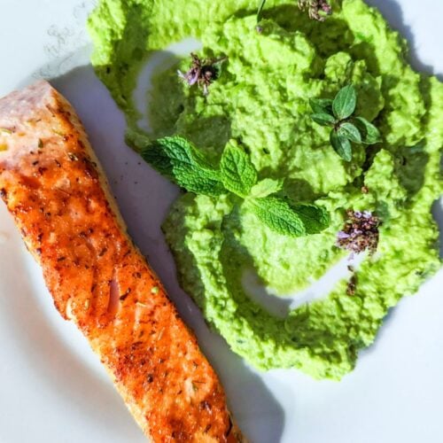 Salmon fillet with crispy edges served alongside smashed peas and fresh mint on a white dinner plate.