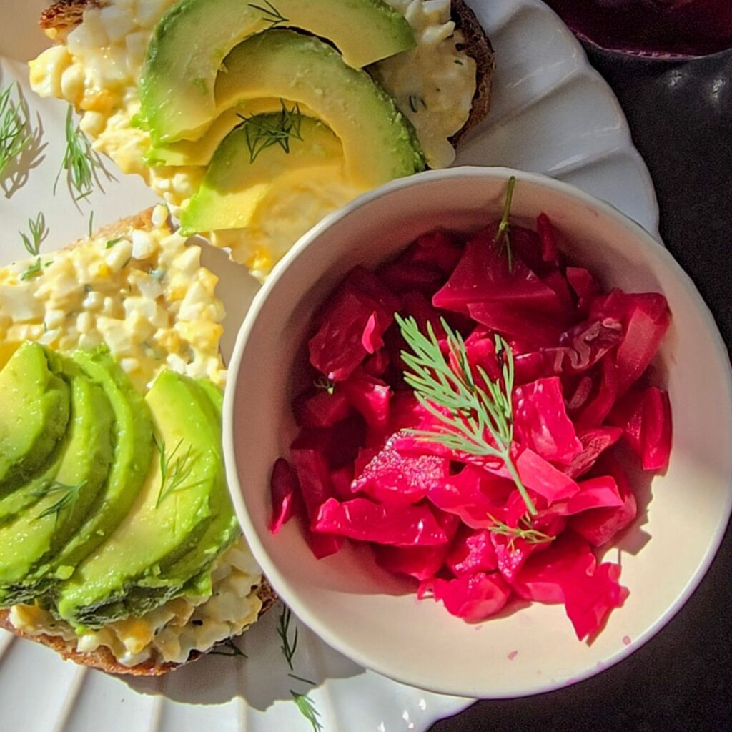 Bowl of pink sauerkraut with dill next to toast topped with egg mayo and sliced avocado.