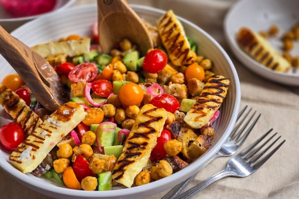 A large bowl of chickpeas and halloumi salad with tomatoes cucumbers and croutons and serving spoons mixing the salad
