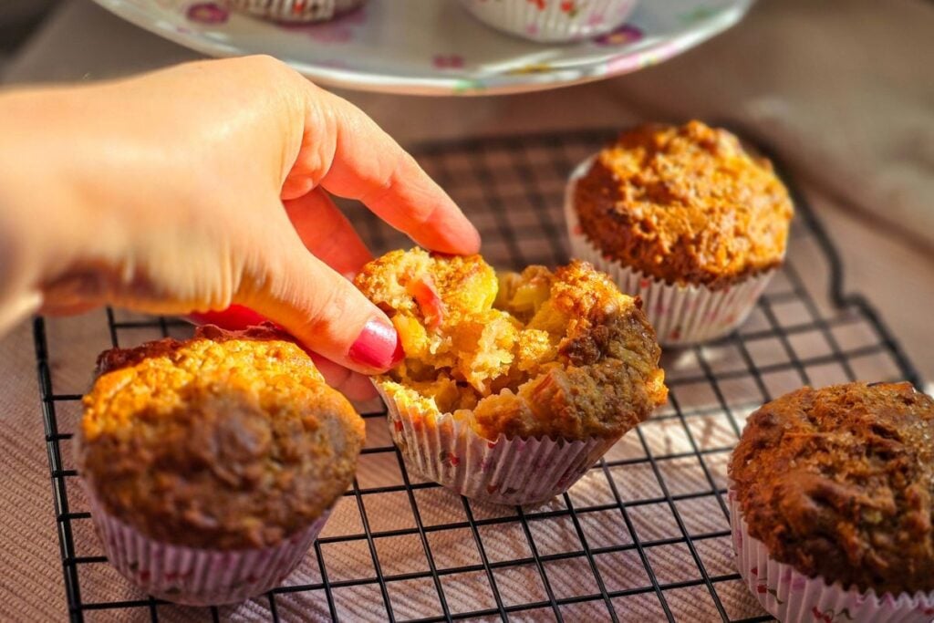 opening up a rhubarb and apple muffin by hand and other muffins on a black cooling grid