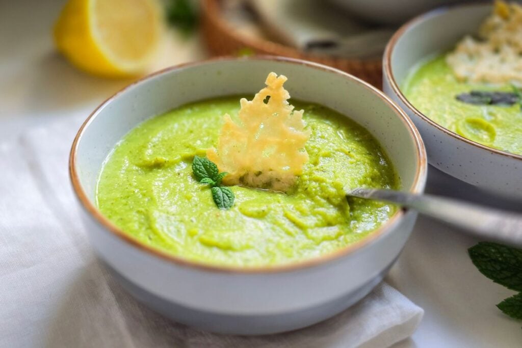 A bowl of bright green courgette and pea soup with a crispy parmesan round on top and half a lemon on the side