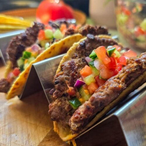 Three smashed burger tacos arranged neatly in taco holders, each filled with a crispy-edged beef patty, melted cheese, and topped with fresh tomato-cucumber salsa. The tortillas are slightly charred and golden, with vibrant colours from the red tomatoes, green cucumber, and fresh herbs, set against a rustic wooden background.