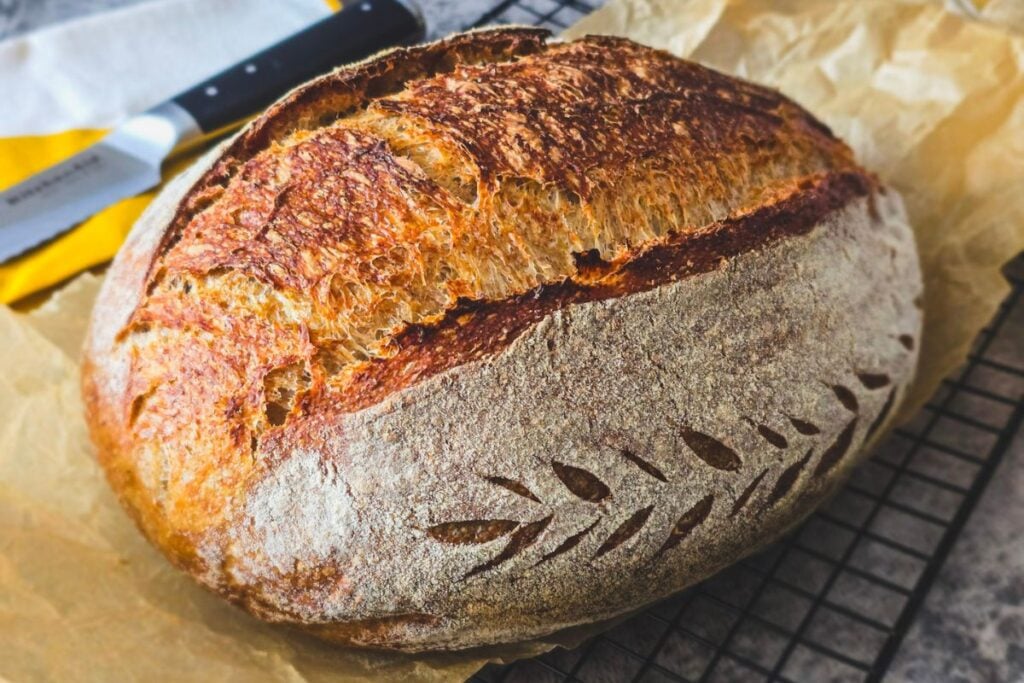 Whole brown sourdough loaf on a cooling rack with deep golden crust and leaf scoring.