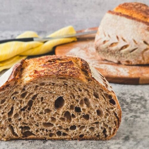 Brown sourdough crumb in foreground with a scored loaf behind, highlighting crust and ear.