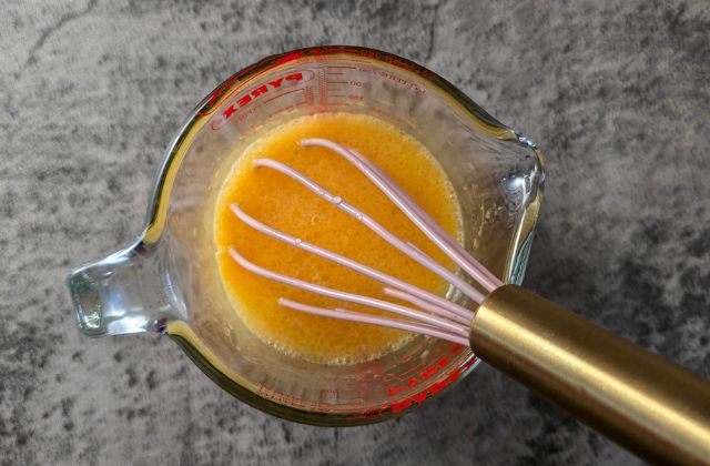Egg yolks mix in glass jug being whisked with pink and gold whisk.