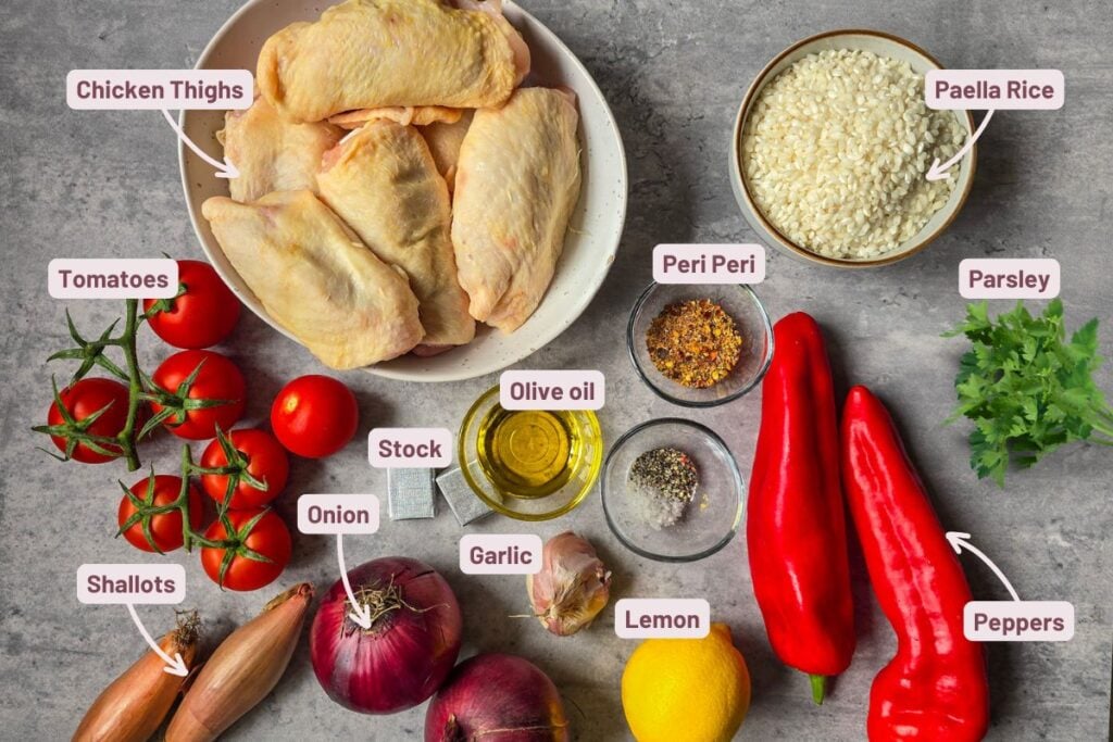 Ingredients for Portuguese-inspired peri peri chicken and rice—chicken thighs, paella rice, red peppers, tomatoes, onions, shallots, garlic, lemon, stock, olive oil, peri-peri seasoning, parsley—labelled on a grey background.