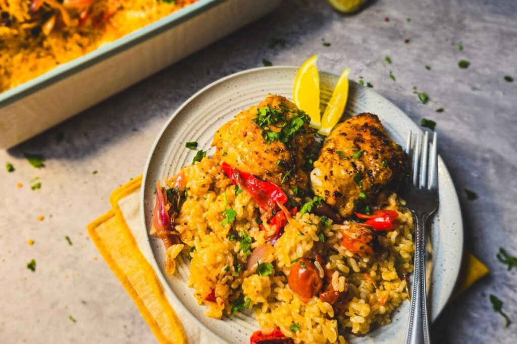 Plate of peri peri chicken thighs over baked rice with peppers and tomatoes, topped with parsley and lemon wedges.