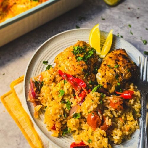 Plate of peri peri chicken thighs over baked rice with peppers and tomatoes, topped with parsley and lemon wedges.