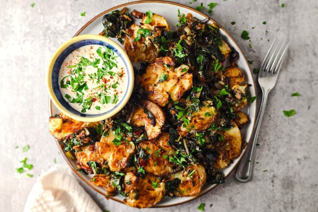 Crispy smashed potatoes with sautéed Swiss chard, parsley, and a bowl of miso-tahini yogurt dressing on a serving platter.