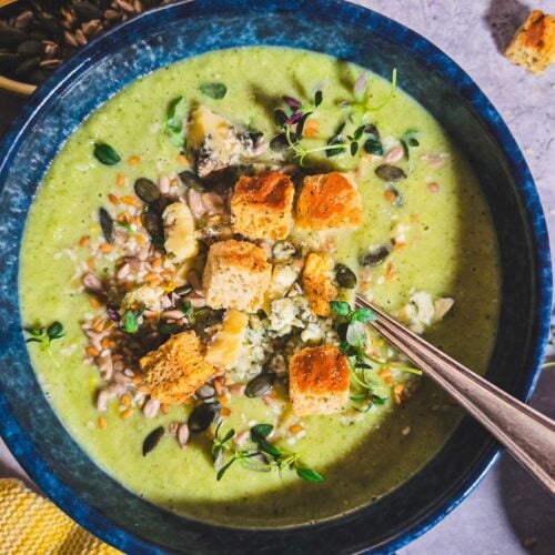 Overhead of broccoli and Stilton soup in a blue bowl, topped with croutons, Stilton crumbles, toasted seeds and thyme, spoon resting inside.