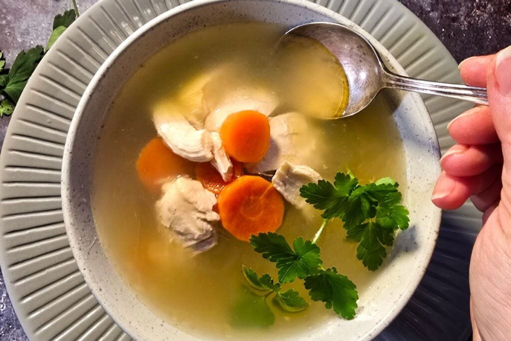 Spoon lifting clear chicken soup with carrots and chicken from a bowl, with parsley on top.