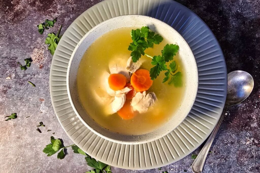 Bowl of clear chicken soup with carrots and chicken pieces, topped with fresh parsley.