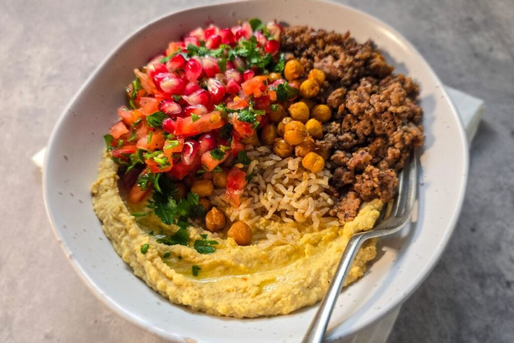 Spiced lamb bowl with hummus, rice, crispy chickpeas, tomato herb salsa, and pomegranate, with a fork resting in the bowl.