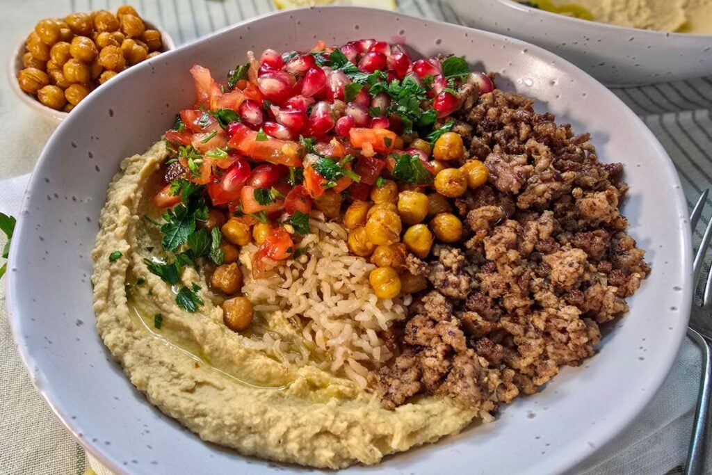 Close-up of a spiced lamb bowl with hummus and crispy chickpeas, topped with tomato herb salsa and pomegranate.