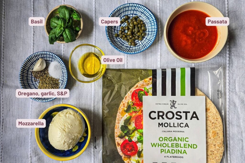 Ingredients for piadina pizza laid out on a table: wholemeal piadina pack, passata, mozzarella, capers, basil, olive oil, and a bowl of oregano, garlic, salt, and pepper.