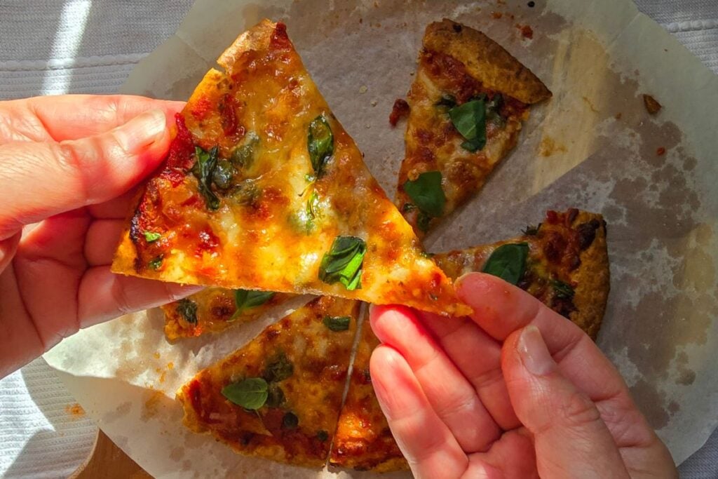 Hand holding a slice of piadina pizza with melted mozzarella and basil over the sliced pizza.