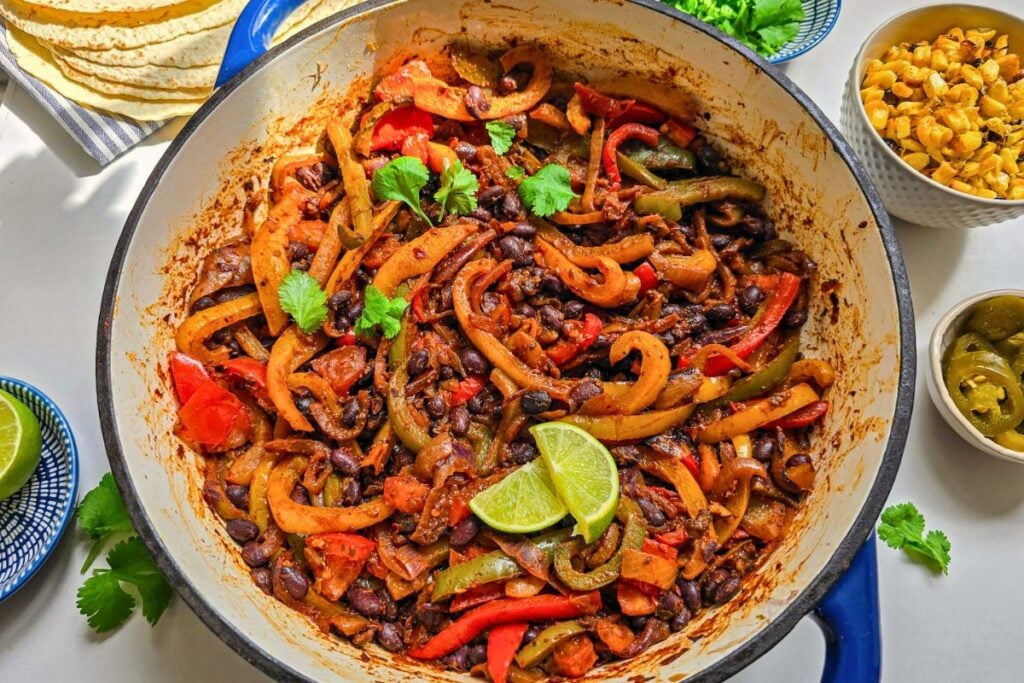 Black bean fajita filling with peppers and onions cooking in a blue skillet, served with lime, jalapeños, corn, herbs, and tortillas.