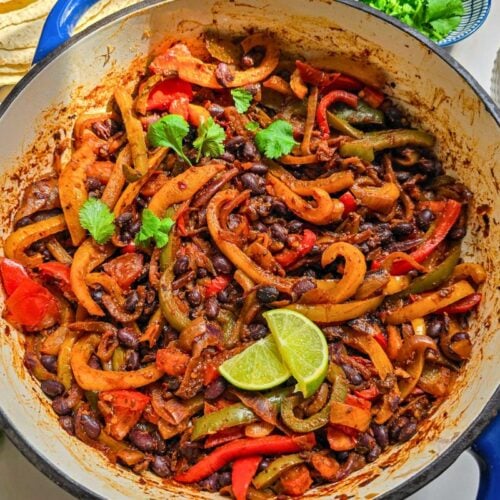 Black bean fajita filling with peppers and onions cooking in a blue skillet, served with lime, jalapeños, corn, herbs, and tortillas.