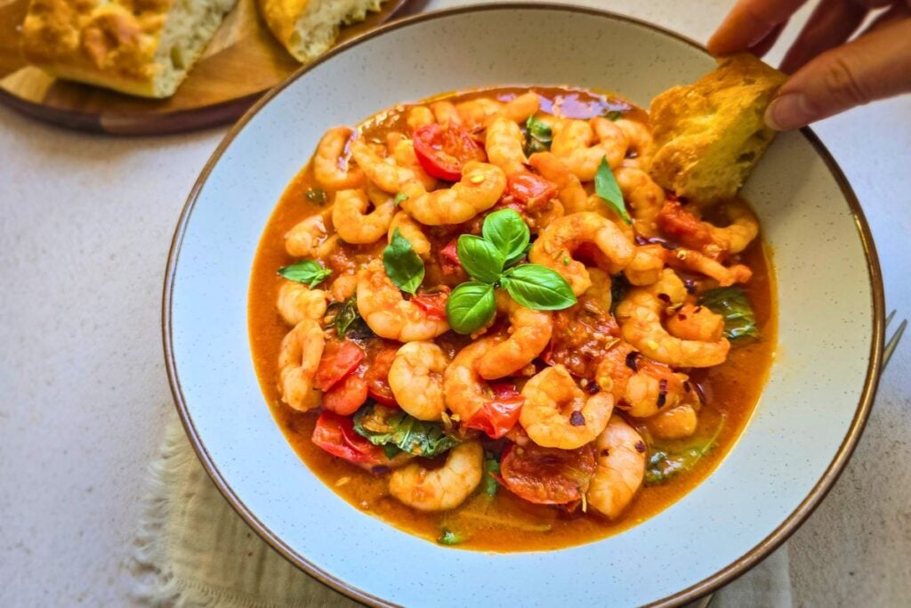 Hand dipping a piece of focaccia into a bowl of shrimp marinara with cherry tomatoes and fresh basil.