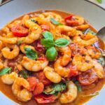 A bowl of saucy shrimp marinara with torn basil leaves and a spoon in.