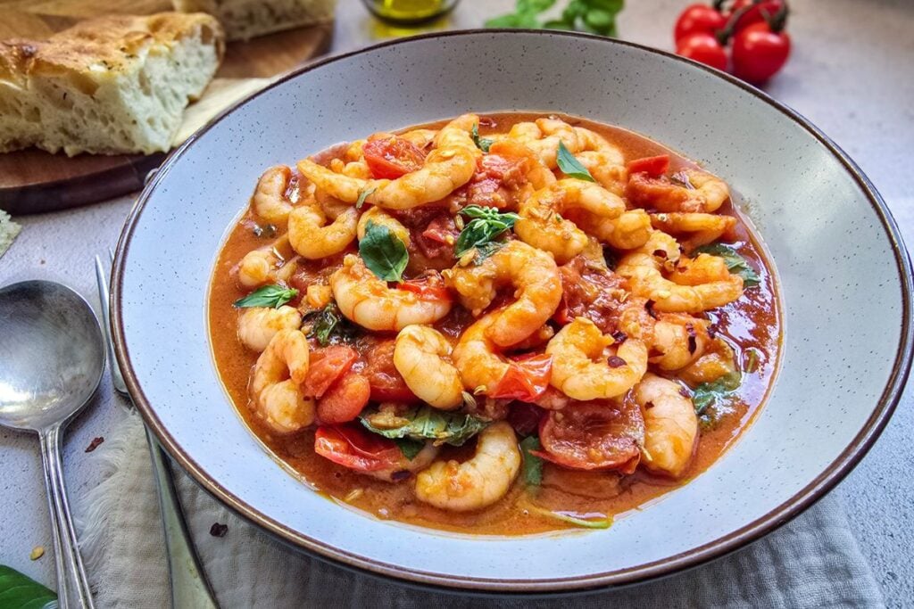 Shrimp marinara in a shallow bowl with cherry tomatoes and basil, served with focaccia on a light linen backdrop.