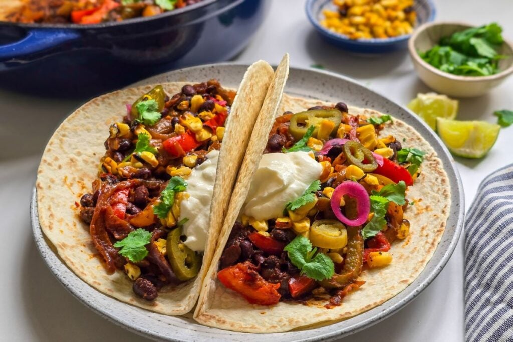 Vegetarian black bean fajitas served in tortillas with corn, pickled onions, jalapeños, cilantro, sour cream, and lime wedges.