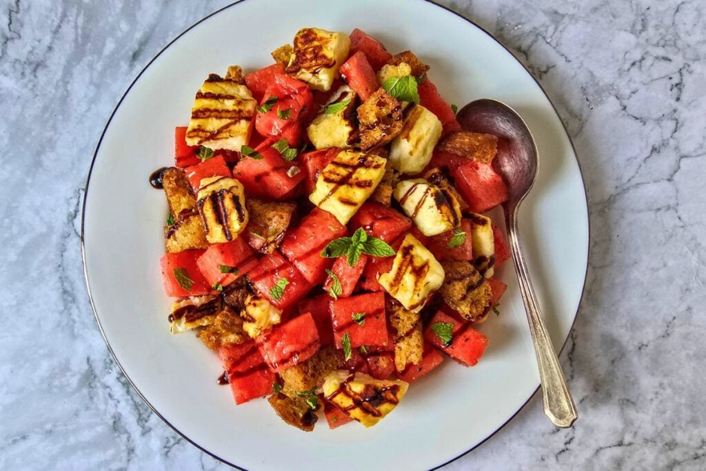 Overhead view of watermelon halloumi salad with grilled sourdough croutons, charred halloumi, fresh mint leaves, and balsamic glaze on a white silver-rimmed plate against a marble surface.