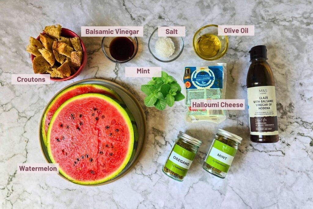 Ingredients for watermelon halloumi salad laid out on a marble surface including a halved watermelon, halloumi, sourdough croutons, balsamic vinegar, balsamic glaze, olive oil, dried oregano, fresh and dried mint, and sea salt.