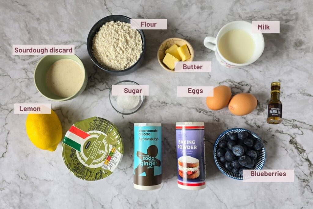 Ingredients for sourdough lemon ricotta pancakes laid out, including ricotta, sourdough discard, flour, eggs, milk, butter, sugar, lemon, vanilla extract, baking powder, bicarbonate of soda, and blueberries