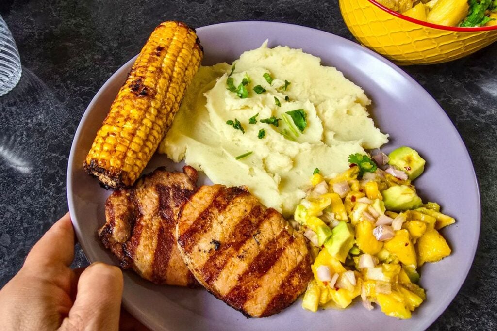 Avocado pineapple salsa served alongside grilled pork chops, mashed potatoes, and charred corn.