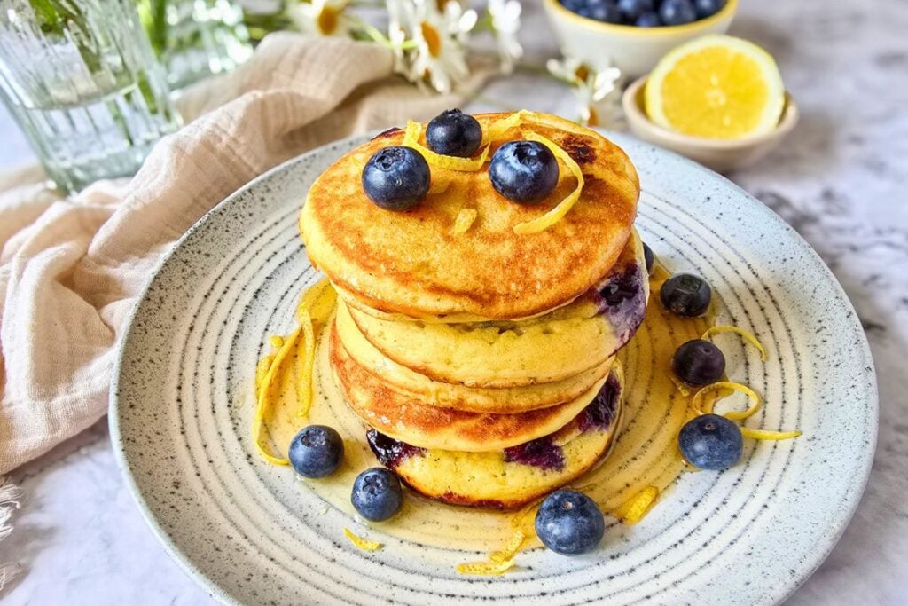 Tall stack of sourdough lemon ricotta pancakes with blueberries, lemon zest curls, and maple syrup on a ceramic plate, with daisies and lemon half in the background