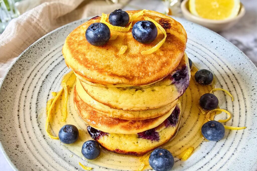 Sourdough lemon ricotta pancakes stacked on a speckled ceramic plate with fresh blueberries, lemon zest curls, and maple syrup