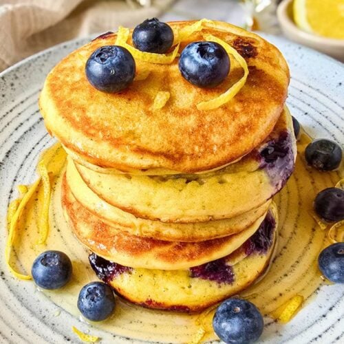 Sourdough lemon ricotta pancakes stacked on a speckled ceramic plate with fresh blueberries, lemon zest curls, and maple syrup