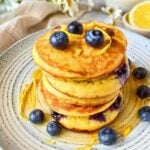 Stack of sourdough lemon ricotta pancakes with blueberries and lemon zest curls on a speckled ceramic plate, with a lemon half and white flowers in the background