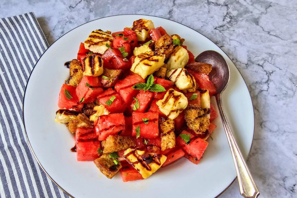 Watermelon halloumi salad with grilled sourdough croutons, fresh mint leaves, and balsamic glaze on a white plate with a silver rim, served on a marble surface.