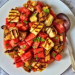 Overhead view of watermelon halloumi salad with charred halloumi, toasted sourdough croutons, fresh mint, and balsamic drizzle on a white plate against a marble background.