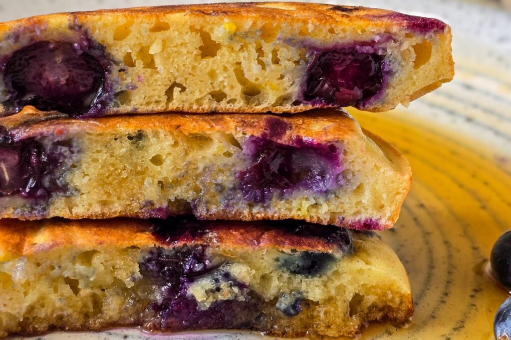 Stack of sourdough lemon ricotta pancakes cut in half to show fluffy interior with blueberries, served with maple syrup on a speckled ceramic plate