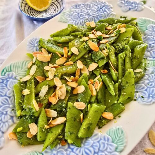 A runner bean salad with gremolata and toasted flaked almonds on a white floral platter and half lemon on the side.