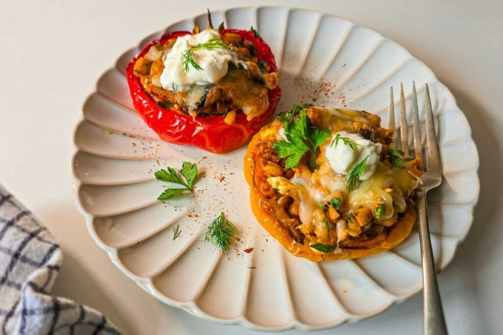 Two barley stuffed pepper halves on a plate, topped with sour cream, dill, and fresh herbs.