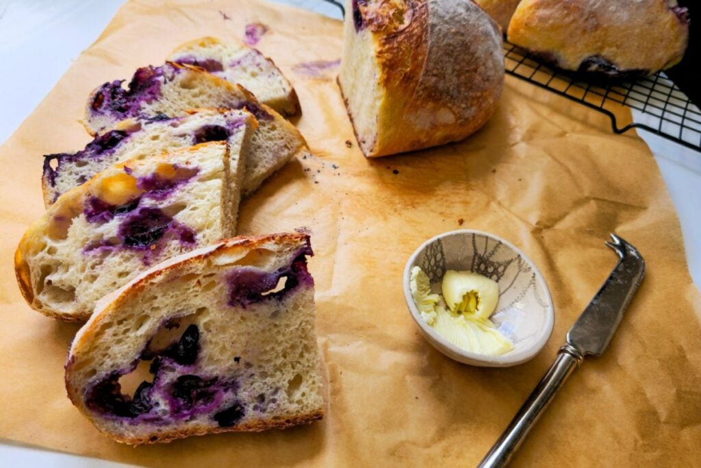 Sliced blueberry sourdough bread showing an open, soft crumb studded with blueberries, next to a small dish of butter and a knife.
