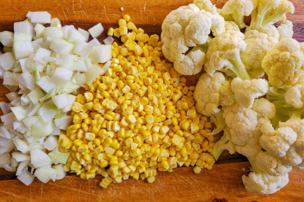 Chopped onion, corn kernels, and cauliflower florets arranged on a wooden board for cauliflower and corn chowder.