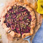 Fig galette with pistachios and balsamic glaze on parchment, with one slice cut and a yellow cloth beside it