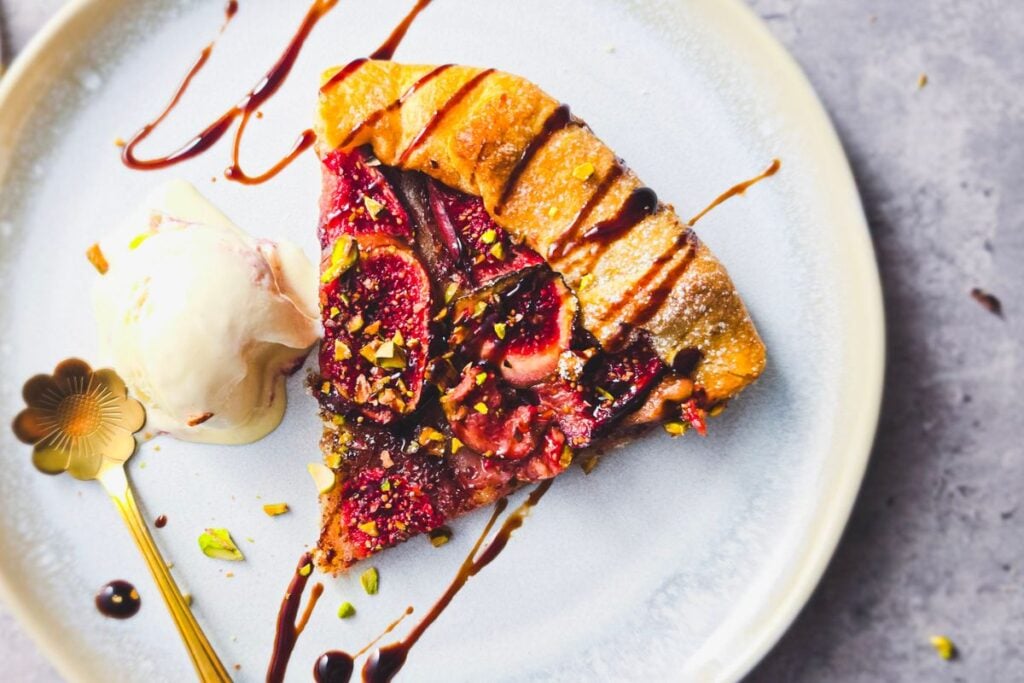 Slice of fig galette with chopped pistachios and balsamic glaze, served with a scoop of ice cream and a gold spoon