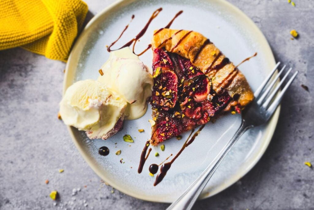 Slice of fig galette on a pale blue plate with two scoops of ice cream, a balsamic glaze drizzle and a fork