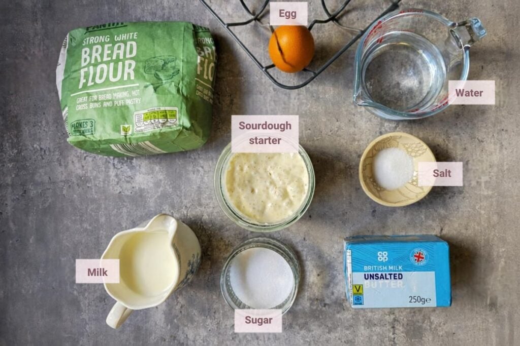 Ingredients for sourdough brioche bread laid out on a gray surface: strong white bread flour, sourdough starter, egg, water, milk, sugar, salt and unsalted butter