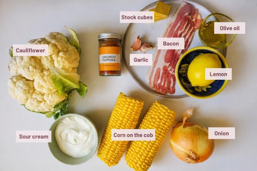 Ingredients for cauliflower and corn chowder: cauliflower, corn on the cob, onion, garlic, bacon, smoked paprika, stock cubes, olive oil, lemon, and sour cream, arranged on a counter.