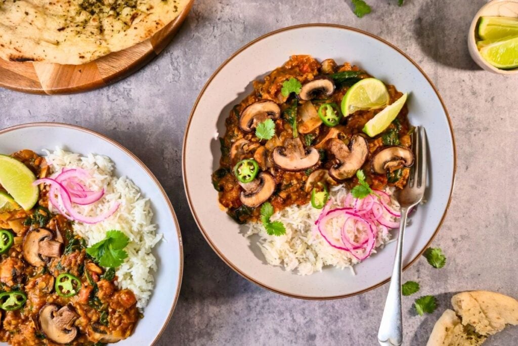 Two bowls of mushroom and spinach curry with rice, lime wedges and garlic naan on a gray table.
