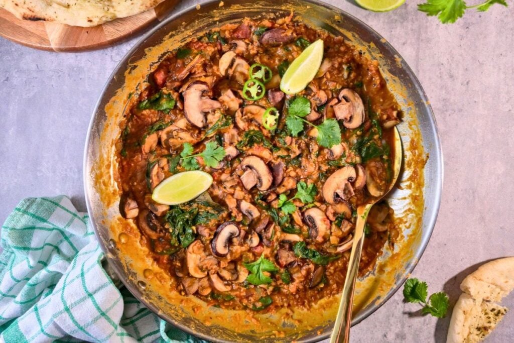 One-pot spinach and mushroom curry with red lentils in a stainless steel pan, topped with cilantro, lime wedges and sliced green chili, with naan on the side.